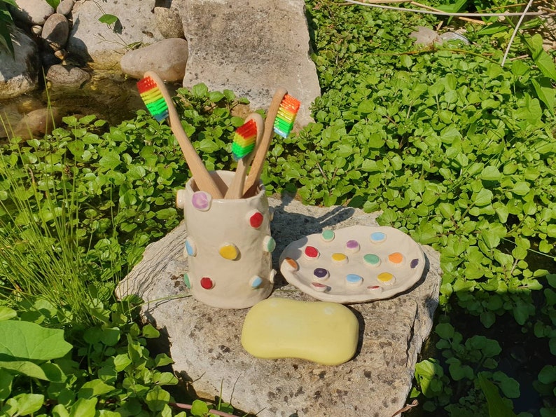 May include: A white ceramic toothbrush holder with colorful polka dots, holding four rainbow-colored toothbrushes. A matching white ceramic dish with colorful polka dots is next to the holder, and a yellow bar of soap is in front of the dish.