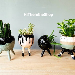 May include: Four ceramic planters with three legs each, painted in black, white, pink, and green with geometric patterns. The planters are filled with various succulents and cacti.