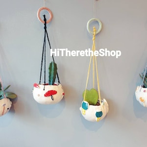 Pode incluir: Quatro vasos suspensos com cordas e designs de cores diferentes. Cada vaso tem uma planta diferente dentro. Os vasos são brancos com designs coloridos. O texto "HiThereTheShop" é visível na imagem.