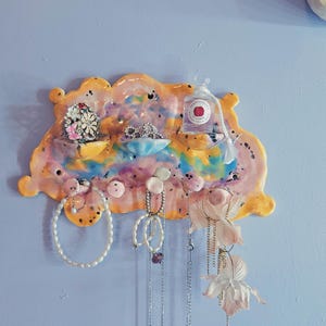 Op de afbeelding: Een kleurrijke, handgemaakte keramische sieradenorganizer in de vorm van een wolk. Het heeft kleine planken en haken voor het uitstallen van sieraden. De organizer is beschilderd met een mix van roze, geel, blauw en oranje tinten en bevat verschillende kettingen, armbanden en oorbellen.