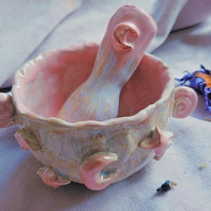 Pode incluir: Um conjunto de almofariz e pilão de cerâmica feito à mão com um esmalte rosa pálido e creme. A taça tem uma borda recortada, duas pegas laterais e decorações em relevo de luas crescentes e estrelas. O pilão também apresenta tons de rosa.