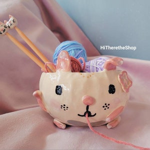 May include: A ceramic yarn bowl shaped like a white bunny with pink ears and a flower on its head. The bowl has a black nose and eyes and is holding two wooden knitting needles with cat-shaped tops. There is blue and purple yarn inside the bowl.