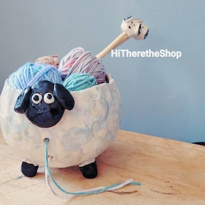 May include: A white ceramic yarn bowl shaped like a sheep with black eyes and a black nose. The bowl is filled with colorful yarn balls and a wooden knitting needle with a white ceramic sheep head on the end. The text "HiThereTheShop" is visible in the background.