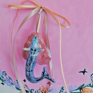 Puede incluir: Un adorno de sirena azul y dorado con una concha rosa y dorada, colgando de una cinta dorada sobre un fondo rosa.