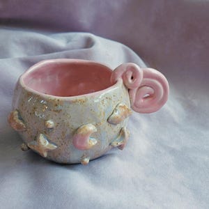 May include: A small, handmade ceramic mug with a pink swirl handle. The mug is decorated with a light blue glaze and has a variety of moon and star shapes.
