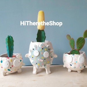May include: Three whimsical ceramic planters, each with a unique design, hold cacti. The planters are white with colorful polka dots and cartoonish faces. One has a tall, yellow cactus, another a green cactus, and the third has a green cactus with flat pads.
