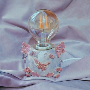 Puede incluir: Una base de lámpara de cerámica con un diseño caprichoso que presenta una luna y flores rosas. La lámpara está iluminada con una bombilla transparente.