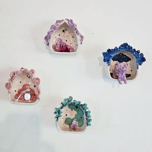 Könnte beinhalten: Vier kleine Keramikhäuser mit unterschiedlich farbigen Dächern und skurrilen Details. Jedes Haus hat eine kleine Figur im Inneren, darunter eine rosa Katze, eine lila Katze, ein grüner Vogel und ein rosa Vogel. Die Häuser sind mit Sternen und Wolken verziert.