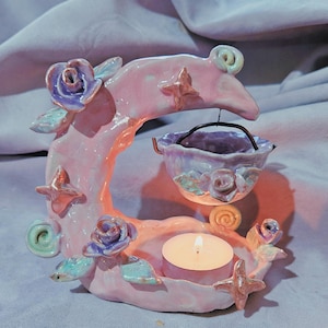 May include: A ceramic crescent moon candle holder in pastel pink with purple rose accents, green leaves, and star shapes. A small bowl hangs from the moon, and a tea light candle sits on the base, creating a soft glow with a glossy finish.