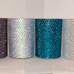 Puede incluir: Siete tazas de diferentes colores cubiertas de piedras de imitación. Las tazas son negras, grises, iridiscentes, azul verdoso, blancas y rosas.