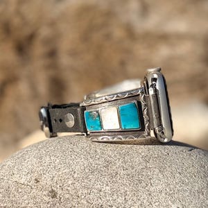 Op de afbeelding: Zilveren Apple Watch-band met een zwarte leren band. De band heeft turkoois en witte stenen inlays. Het horloge ligt op een grijze steen, met een vage achtergrond.