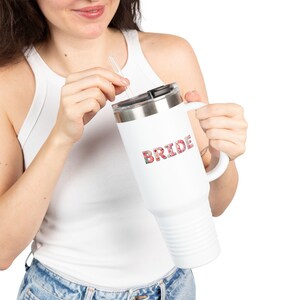 Op de afbeelding: Witte reismok met een handvat en het woord "BRIDE" in een bloemenpatroon. De mok heeft een zilveren deksel en een doorzichtig rietje. De mok wordt vastgehouden door een persoon die een witte tanktop en een blauwe jeans draagt.