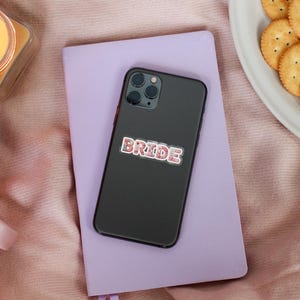 May include: A black smartphone with a floral "BRIDE" sticker rests on a purple notebook. A lit candle in a glass container, a pink mug with tea, and a plate of crackers are also visible.