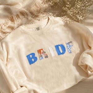 May include: A cream-colored crewneck sweatshirt featuring the word BRIDE appliqued across the chest. The letters are crafted from various blue and white patterned fabrics with visible stitching around the edges of each letter.