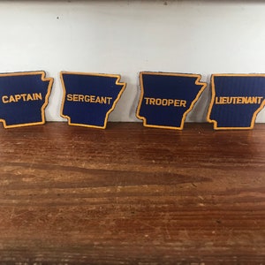 May include: Four blue and gold embroidered patches shaped like the state of Arkansas. The patches read "Captain", "Sergeant", "Trooper", and "Lieutenant".