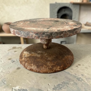 May include: A circular pottery wheel with a weathered, dark brown and rust-colored finish. The flat, rotating top platform is supported by a central pillar attached to a heavy, dome-shaped base, showing signs of oxidation and age.