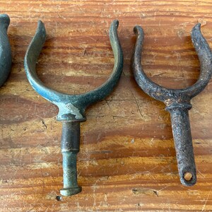 May include: Four antique metal boat cleats with a weathered finish. The cleats are in the shape of a U and have a pointed end. They are made of a dark metal with a green patina.