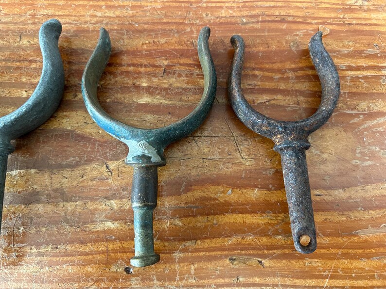 May include: Four antique metal boat cleats with a weathered finish. The cleats are in the shape of a U and have a pointed end. They are made of a dark metal with a green patina.
