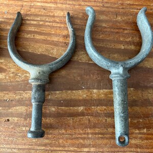 May include: Four antique metal boat cleats with a u-shaped hook and a long, round post. The cleats are weathered and have a dark gray finish.