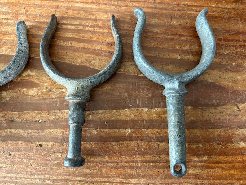 May include: Four antique metal boat cleats with a u-shaped hook and a long, round post. The cleats are weathered and have a dark gray finish.