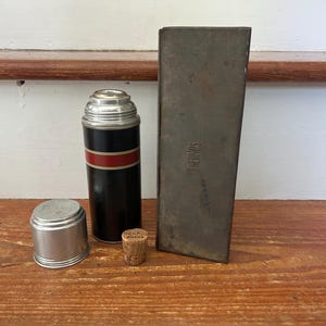 May include: A vintage Thermos brand vacuum flask with a black body, a red and white horizontal stripe, and a silver metal cap. It includes a separate silver metal cup, a small cork stopper labelled Thermos Brand, and a tall, rectangular metal case.