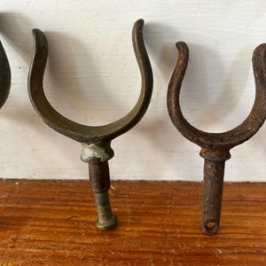May include: Three antique metal boat hooks with a U-shaped design. The hooks are made of different metals, with one being a lighter shade of metal and the other two being a darker shade of metal.
