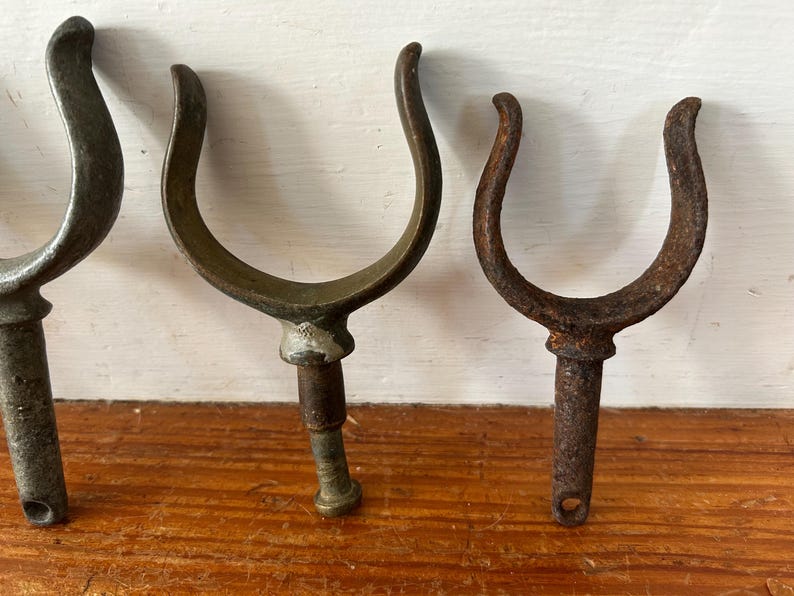 May include: Three antique metal boat hooks with a U-shaped design. The hooks are made of different metals, with one being a lighter shade of metal and the other two being a darker shade of metal.