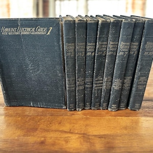 以下が含まれることがあります： 金色の文字で「Hawkins Electrical Guide」と題されたヴィンテージ本のコレクション。本は濃い青と黒で、木製の表面に配置されています。背表紙が見え、巻数が表示されています。