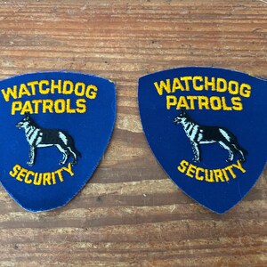 May include: Two blue patches with yellow embroidered text that reads "WATCHDOG PATROLS SECURITY". Each patch features a black and white dog silhouette. The patches are shield-shaped.