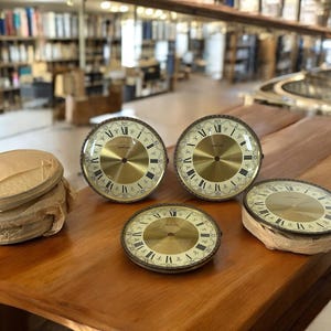 Puede incluir: Varios cuadrantes de reloj vintage con esferas doradas y números romanos negros se exhiben sobre una superficie de madera. Los cuadrantes están en varias etapas de embalaje. El fondo muestra un entorno de biblioteca.