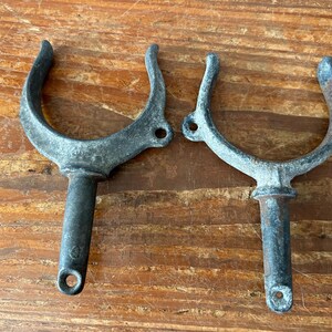 May include: Two weathered metal boat cleat hooks with a U-shaped design and a long, cylindrical stem. The hooks are made of a dark gray metal and have a rustic appearance.