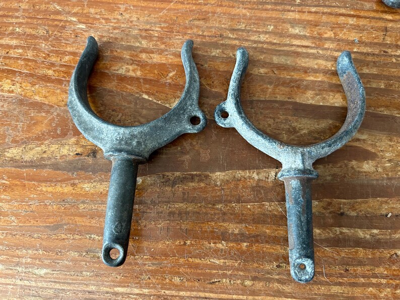 May include: Two weathered metal boat cleat hooks with a U-shaped design and a long, cylindrical stem. The hooks are made of a dark gray metal and have a rustic appearance.