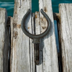 May include: A black metal boat cleat, shaped like a horseshoe, is mounted on a weathered wooden dock. The cleat is used to secure a boat to the dock.