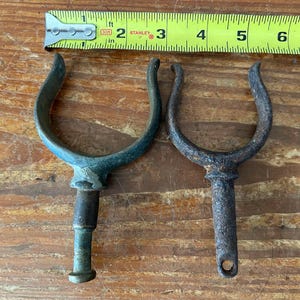 May include: Two vintage boat cleat hooks. One is green and the other is rusty brown. Both are made of metal and have a U-shaped design.