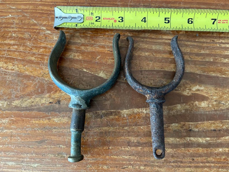 May include: Two vintage boat cleat hooks. One is green and the other is rusty brown. Both are made of metal and have a U-shaped design.