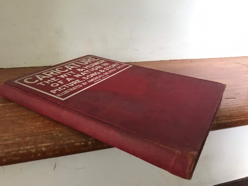 May include: A red book titled "Caricature: The Wit & Humor of a Nation in Picture, Song & Story" with silver lettering. The book is illustrated by America's Greatest Artists.