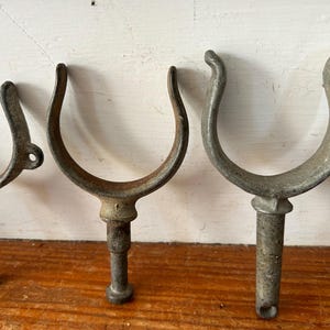 May include: Five antique metal boat cleat hooks with a U-shaped design and a threaded post. The hooks are made of a weathered metal with a gray and brown patina.