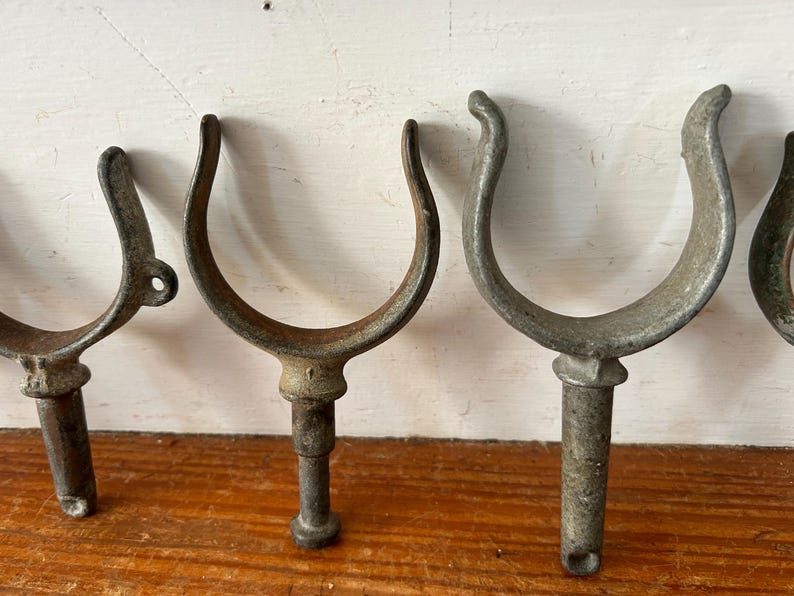 May include: Five antique metal boat cleat hooks with a U-shaped design and a threaded post. The hooks are made of a weathered metal with a gray and brown patina.