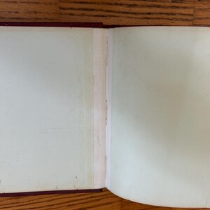 Pode incluir: Um livro aberto com p&aacute;ginas esbranqui&ccedil;adas, mostrando sinais de idade. A lombada &eacute; preta e as bordas da capa s&atilde;o vermelho escuro. As p&aacute;ginas apresentam ligeira descolora&ccedil;&atilde;o e imperfei&ccedil;&otilde;es, sugerindo um item antigo.