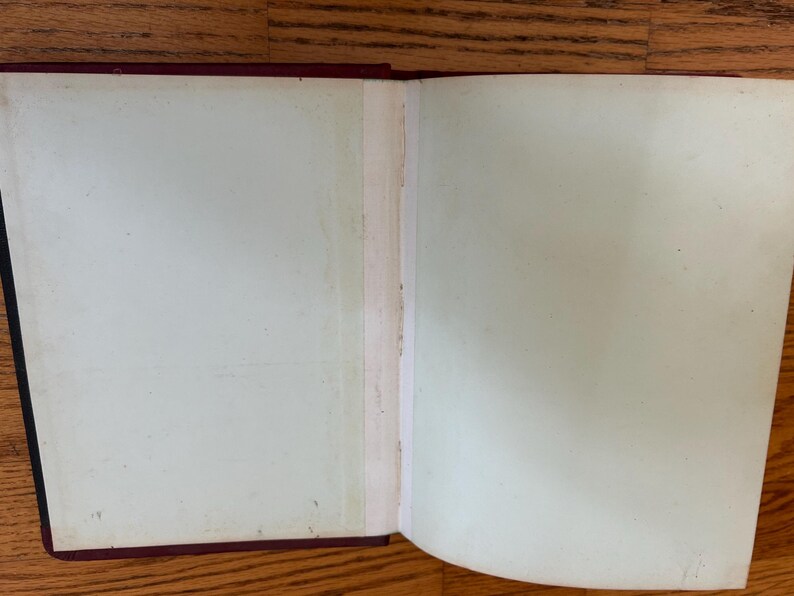 Pode incluir: Um livro aberto com p&aacute;ginas esbranqui&ccedil;adas, mostrando sinais de idade. A lombada &eacute; preta e as bordas da capa s&atilde;o vermelho escuro. As p&aacute;ginas apresentam ligeira descolora&ccedil;&atilde;o e imperfei&ccedil;&otilde;es, sugerindo um item antigo.
