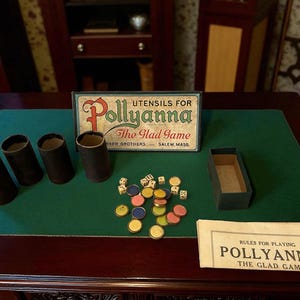 Op de afbeelding: Een antieke Pollyanna bordspelset met een groene speeloppervlak. De set bevat een doos, dobbelstenen, gekleurde fiches en vier bruine papieren cilinders. De speldoos leest "Pollyanna The Glad Game". Er is ook een regelvel aanwezig.
