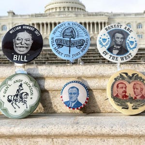 May include: A collection of vintage political campaign buttons featuring portraits of historical figures. The buttons are round and colorful, with text and images. The background includes the U.S. Capitol building.