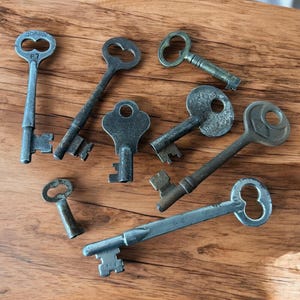 May include: A collection of antique keys in various sizes and designs, made of metal. The keys are arranged on a wooden surface, showcasing their intricate details and aged appearance. Some keys have markings.