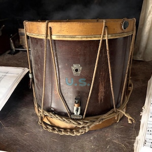 Puede incluir: Un tambor de madera vintage con acabado marrón oscuro y detalles en marrón claro. El tambor presenta las letras "U.S." en azul y un escudo. Se utilizan cuerdas y cordones para la construcción del tambor. Partituras visibles en el fondo.