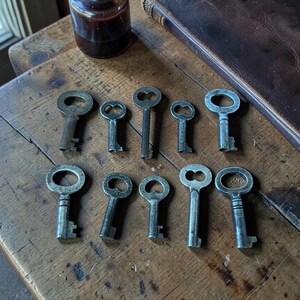 May include: A collection of antique metal keys of various sizes and designs arranged on a weathered wooden surface. A dark leather-bound book and a small glass bottle are also present. The keys have ornate keyholes and stems.