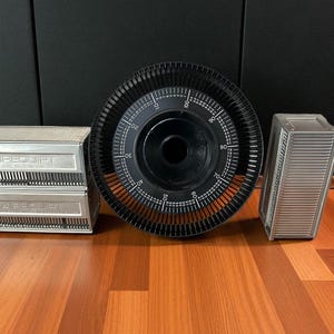 May include: A collection of vintage slide projectors and accessories. Two silver metal slide trays are stacked, with the word "AIREQUIP" printed on them. A large black slide carousel and a silver metal slide holder are also present.
