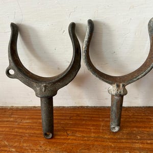 May include: Two vintage metal boat cleat hooks with a U-shaped design. The hooks are made of dark gray metal and have a rusty patina. The hooks are attached to a metal rod.