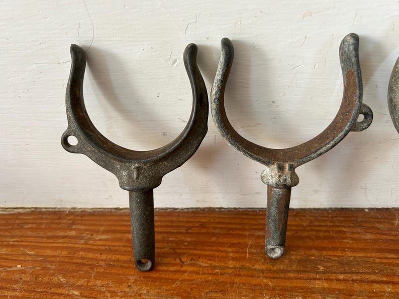 May include: Two vintage metal boat cleat hooks with a U-shaped design. The hooks are made of dark gray metal and have a rusty patina. The hooks are attached to a metal rod.