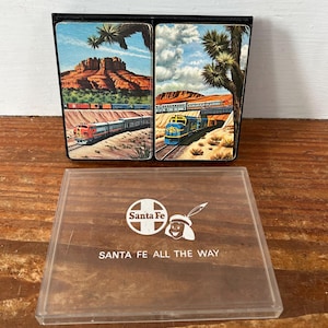 Puede incluir: Una taza de cerámica blanca con un asa negra y el texto "But First, Coffee" en negrita negra. La taza se encuentra sobre un fondo borroso, sugiriendo un ambiente acogedor.