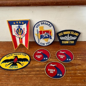May include: A collection of six embroidered patches featuring various military and aircraft designs. The patches are yellow, red, blue, and white. The patches include the names "USS Ohio", "USS Dallas", "USS Rhode Island", "Sam Houston", and "Eagle 100".